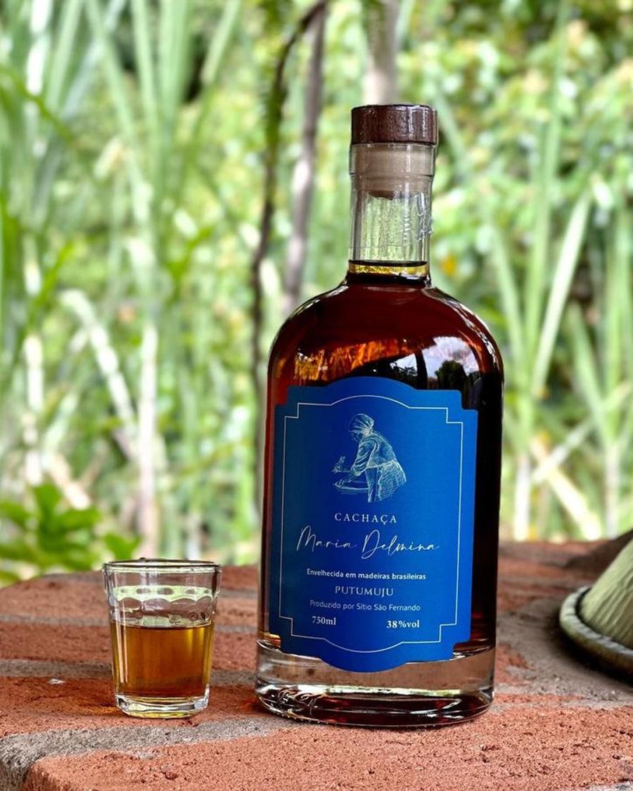 Garrafa da cachaça Maria Delmina Putumuju ao lado de um copinho, com canavial ao fundo