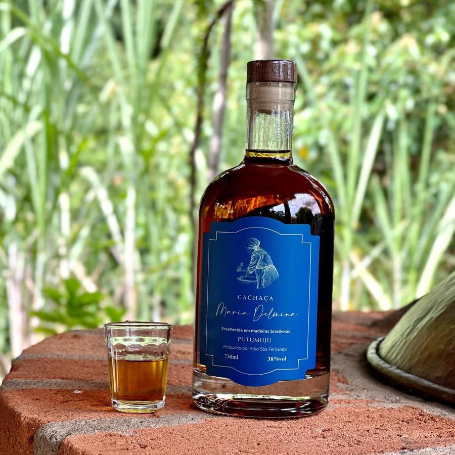 Garrafa da cachaça Maria Delmina Putumuju ao lado de um copinho, com canavial ao fundo